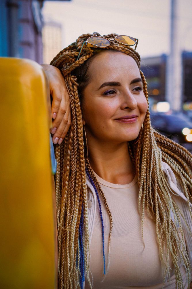 Young person with braided hairstyle, some braids pulled up, wearing a black salon cape, indoors.