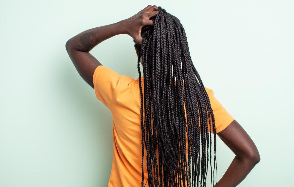Young person with braided hairstyle, some braids pulled up, wearing a black salon cape, indoors.
