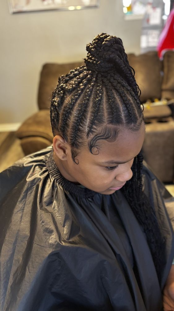 Young person with braided hairstyle, some braids pulled up, wearing a black salon cape, indoors.