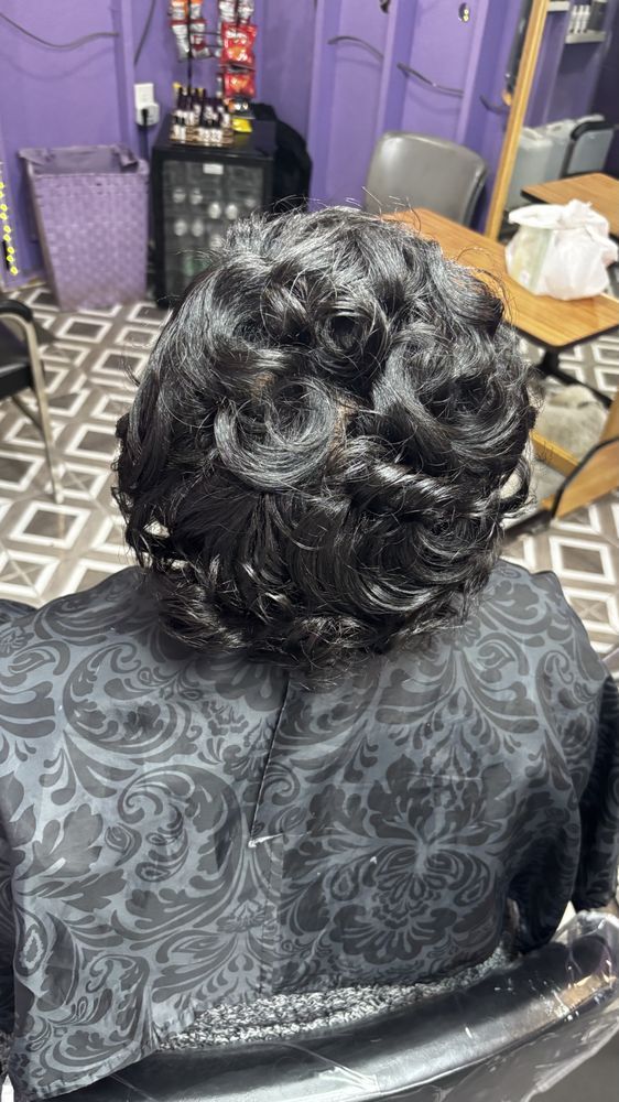 Back view of a person with short, curly black hair. They wear a black patterned top. Indoors, salon setting.