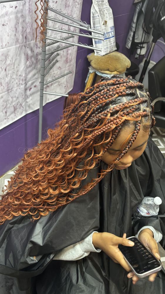 Woman with orange braided hair, looking down at a phone, in a salon setting.