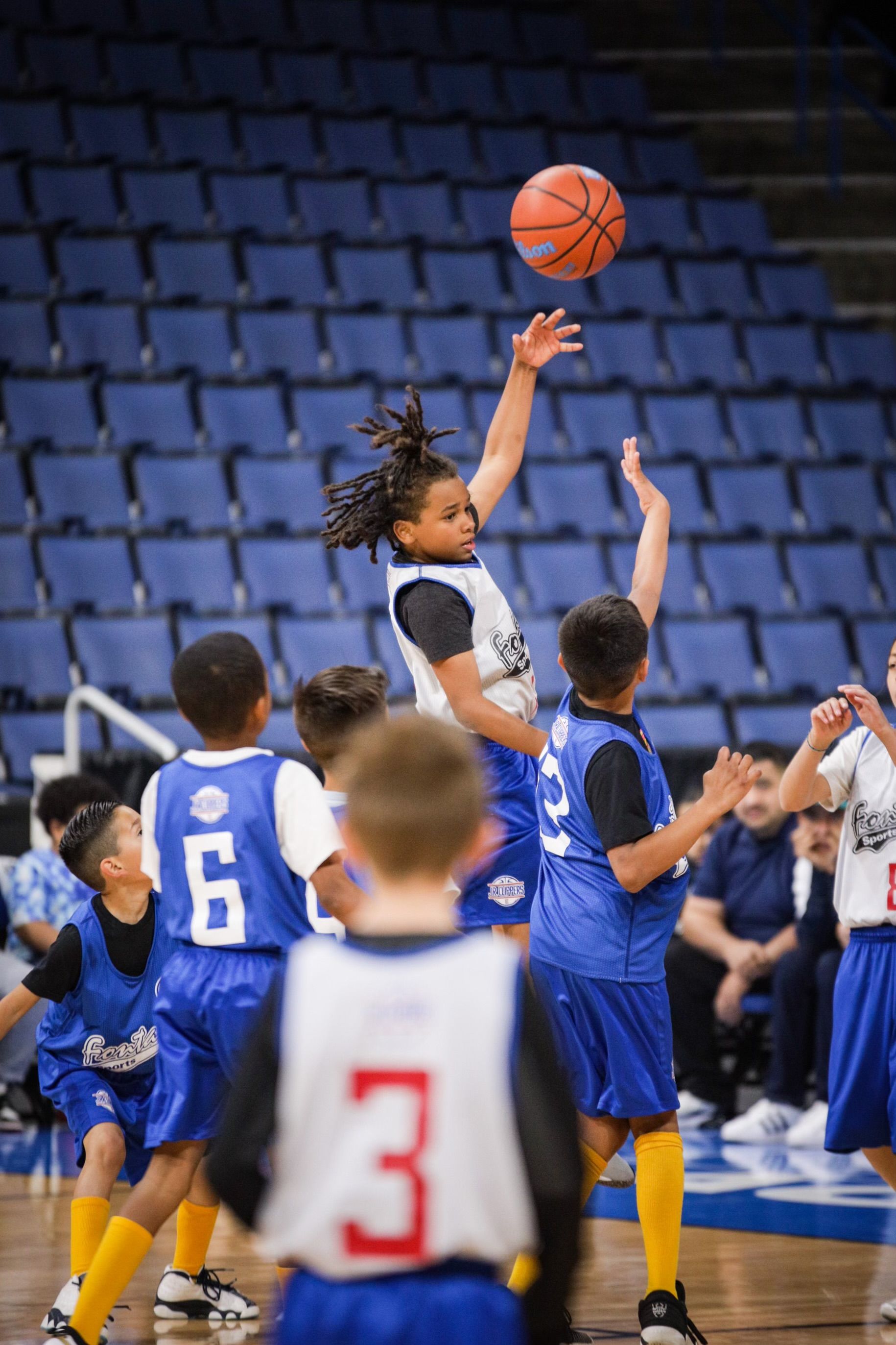 Youth basketball players in blue jerseys compete for the ball on a court inside an arena with blue stadium seating.