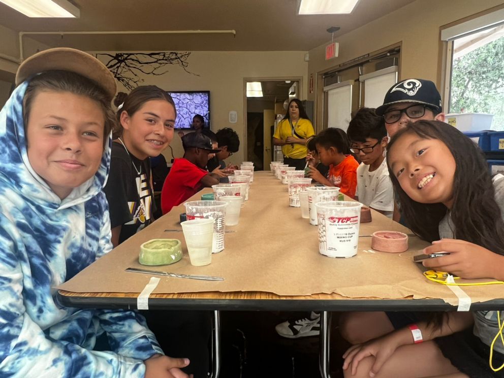 A group of students sits at a long table during a craft activity with supplies and containers in a room.