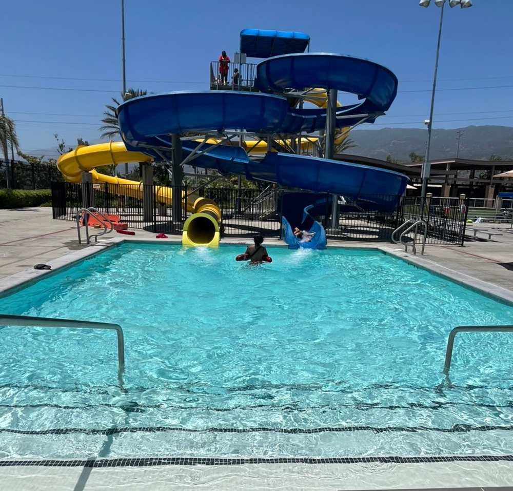 A swimming pool with two large, winding slides in blue and yellow, with people playing in the water on a sunny day.