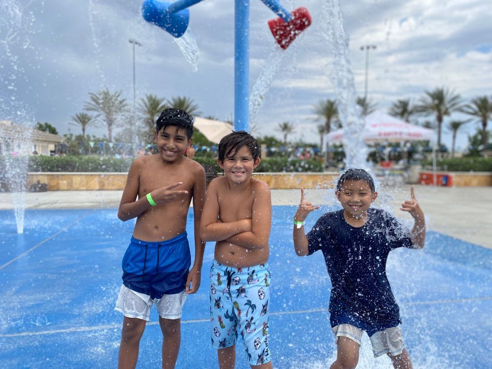 Three children smile while playing in water sprinklers at an outdoor splash pad on a cloudy day.