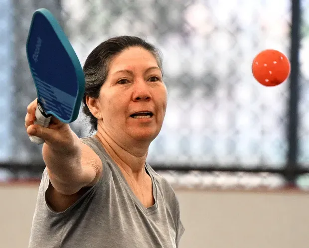 A person in a gray shirt holding a blue pickleball paddle, ready to hit an orange ball on a court.