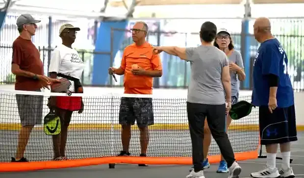 A group of people standing on a pickleball court, talking near the net while holding paddles.