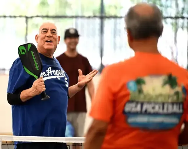 Three people play pickleball indoors; a man in a blue Los Angeles shirt holds a green paddle while talking to a partner.