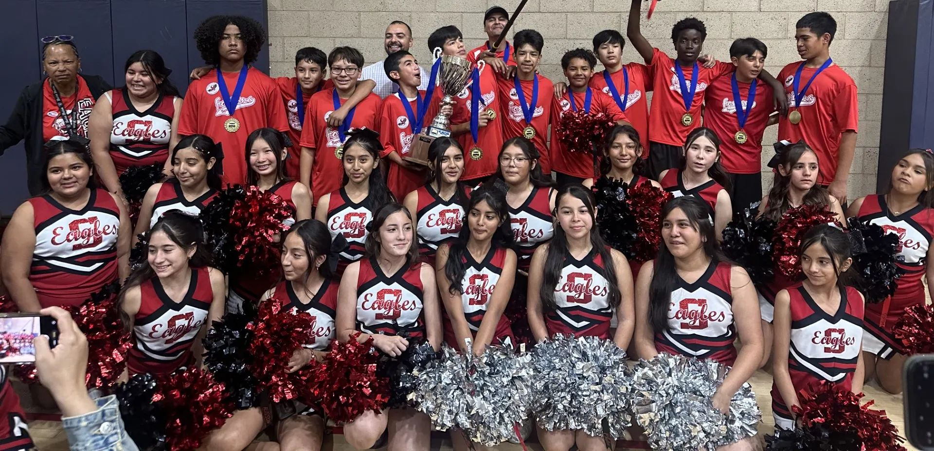 A large team of cheerleaders and athletes pose together indoors, many holding medals and a trophy, wearing red uniforms.