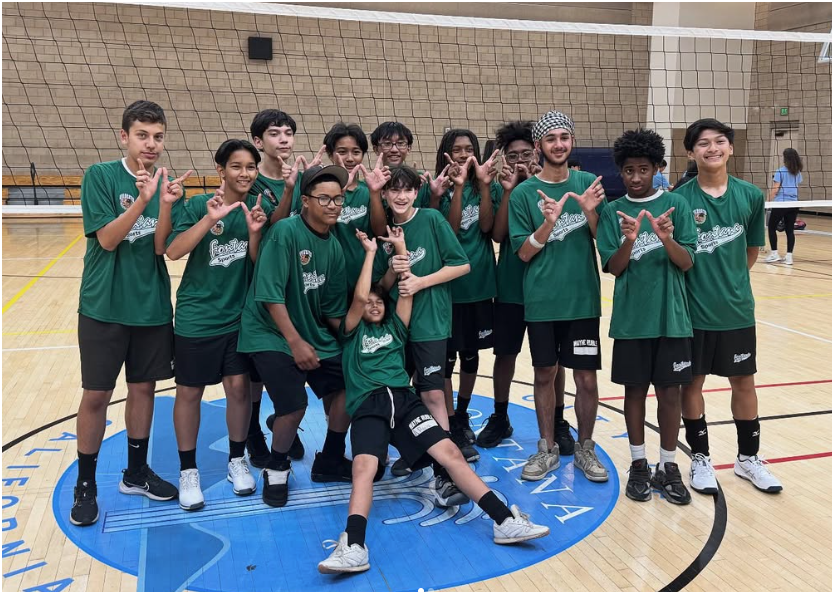 A team of thirteen people in matching green shirts and black shorts stand in a gym, posing with hand signs on a court.