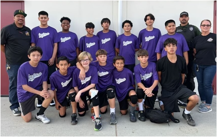 A team in matching purple t-shirts poses for a group photo outdoors with two coaches on a concrete surface.