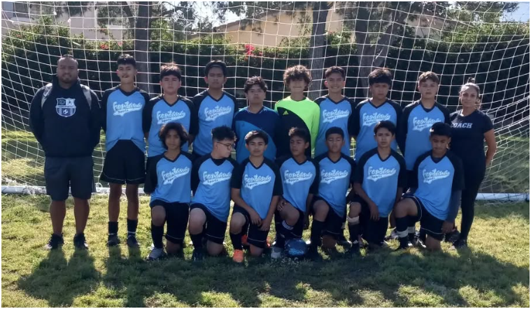 A youth soccer team in blue jerseys and black shorts poses for a group photo in front of a goal net on a grassy field.