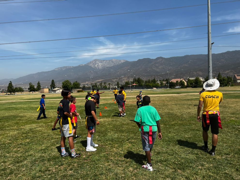 A coach in a yellow shirt stands with a youth flag football team on a grassy field against a backdrop of mountains.