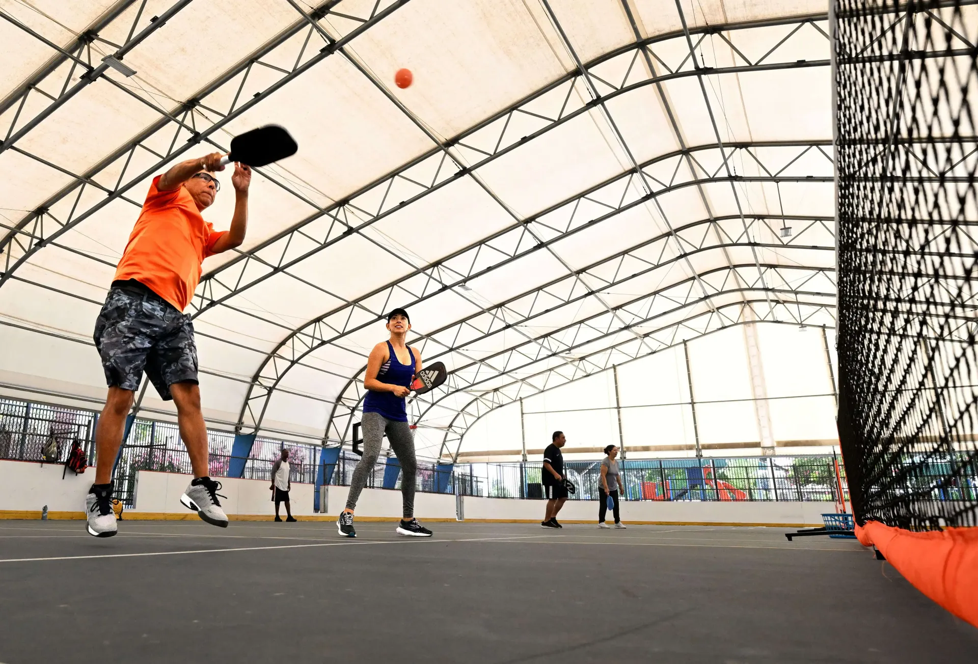 People playing pickleball on an indoor court under a high, arched frame structure.