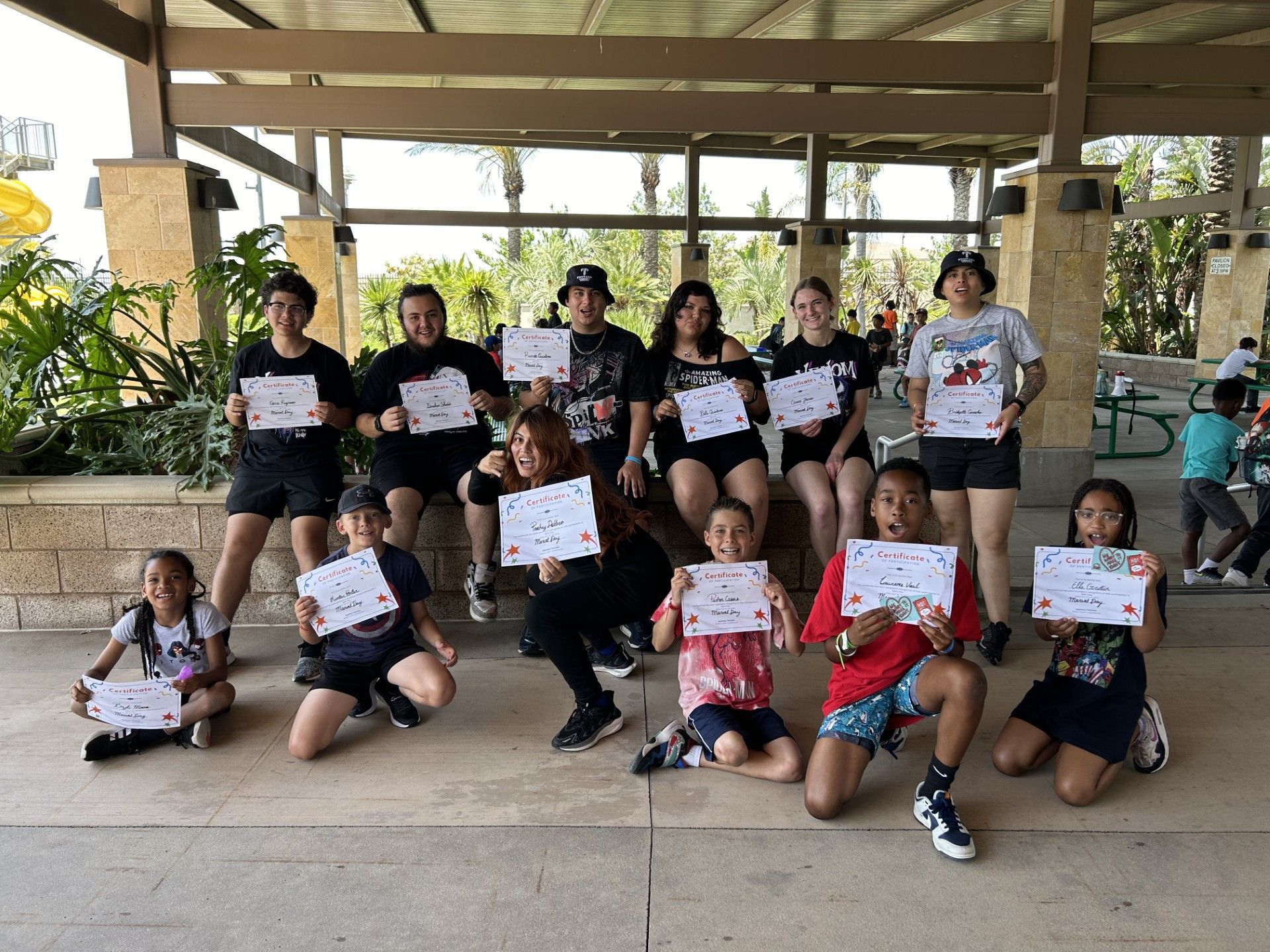 A group of people holding certificates, posing together outdoors under a shaded pavilion.