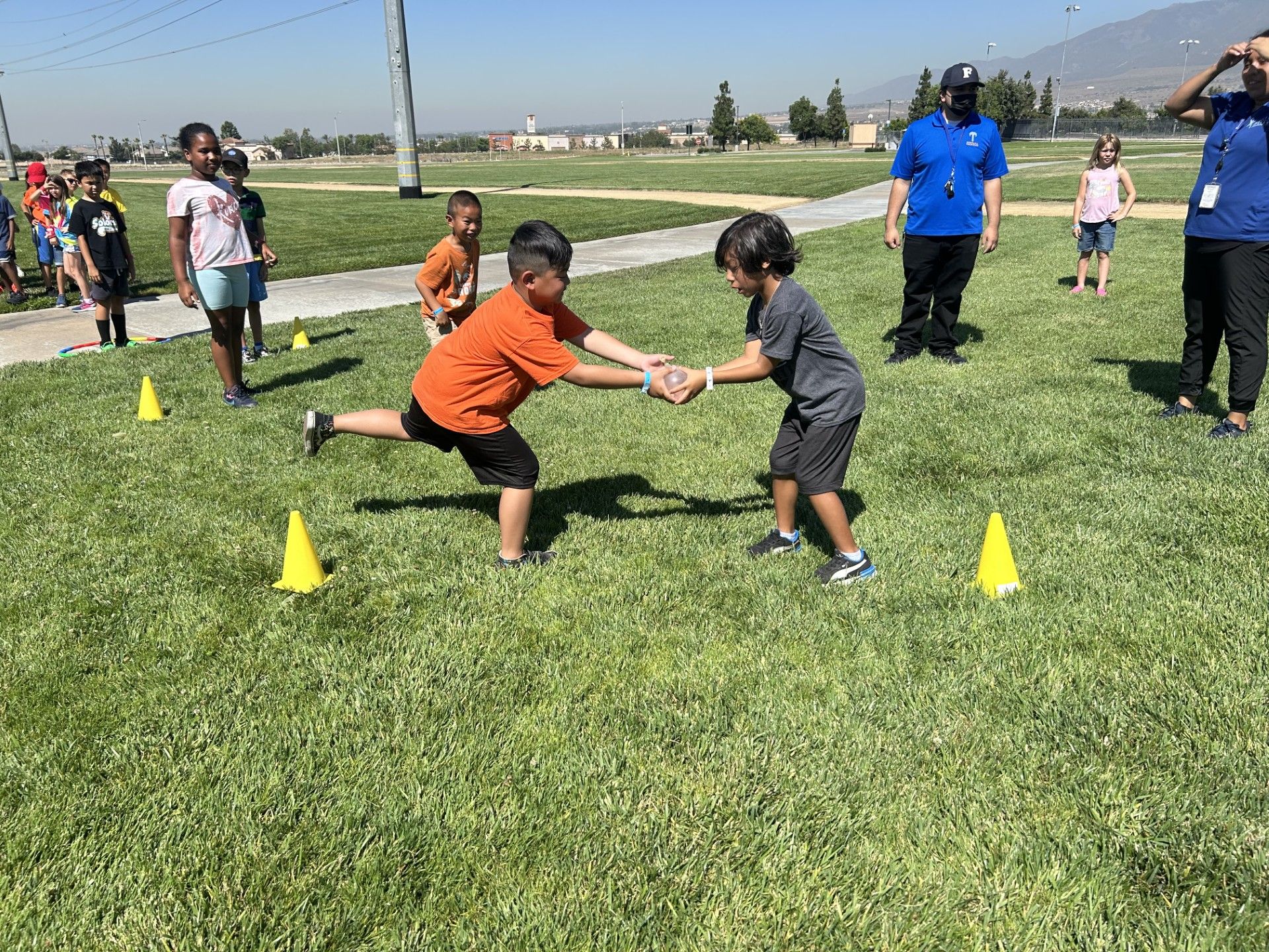 Two children hold hands while balancing on one leg during an outdoor physical activity on a sunny, grassy field.