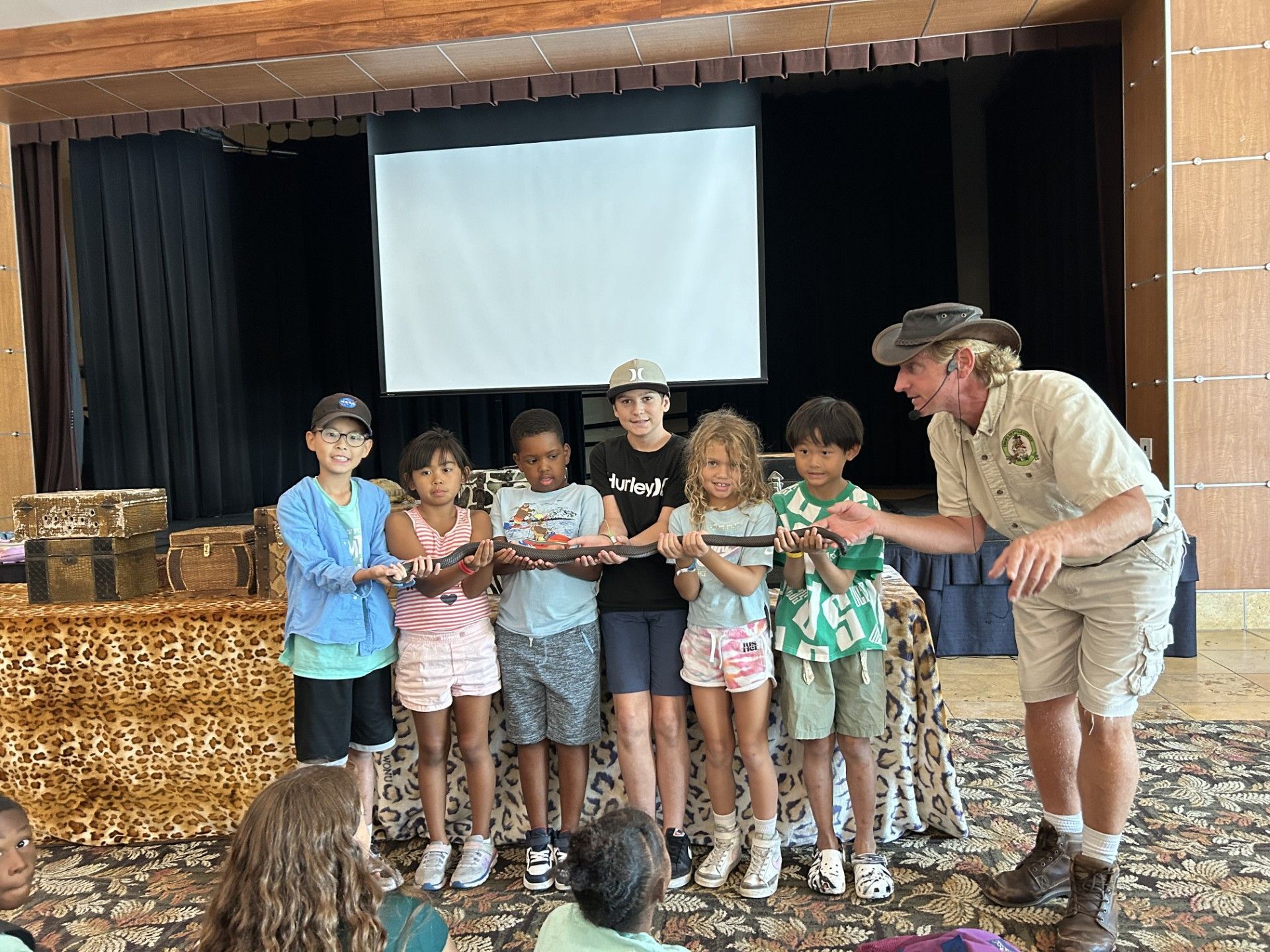A group of children watch as a person in safari gear holds a small animal for them to touch during an educational event.
