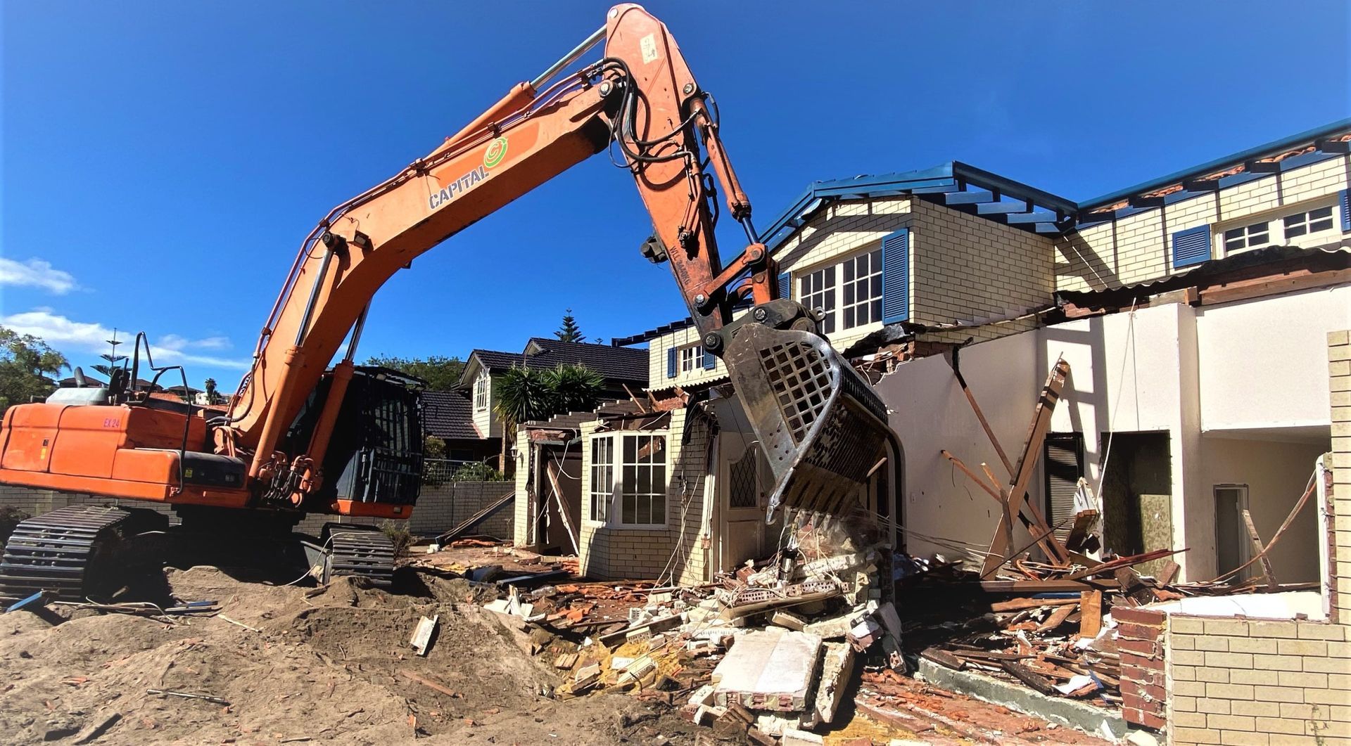 Excavator demolishing a damaged house, with rubble and debris scattered under a blue sky.