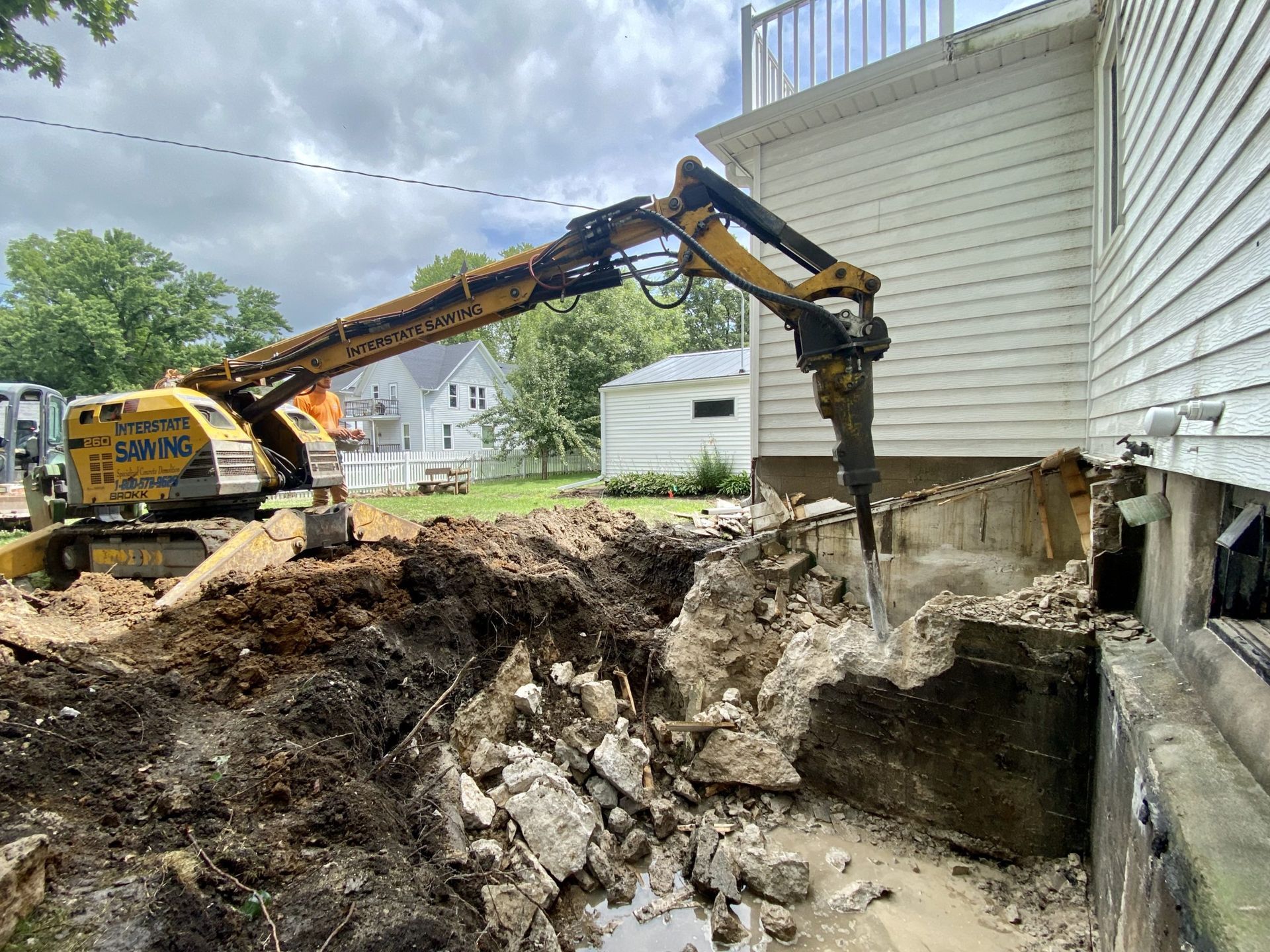 Excavator digging beside a house, removing dirt and concrete near the foundation
