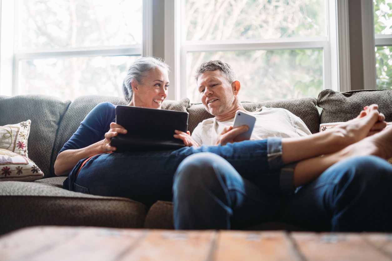 Two people sitting together on a couch, looking at a tablet and a smartphone in a bright, relaxed home setting.