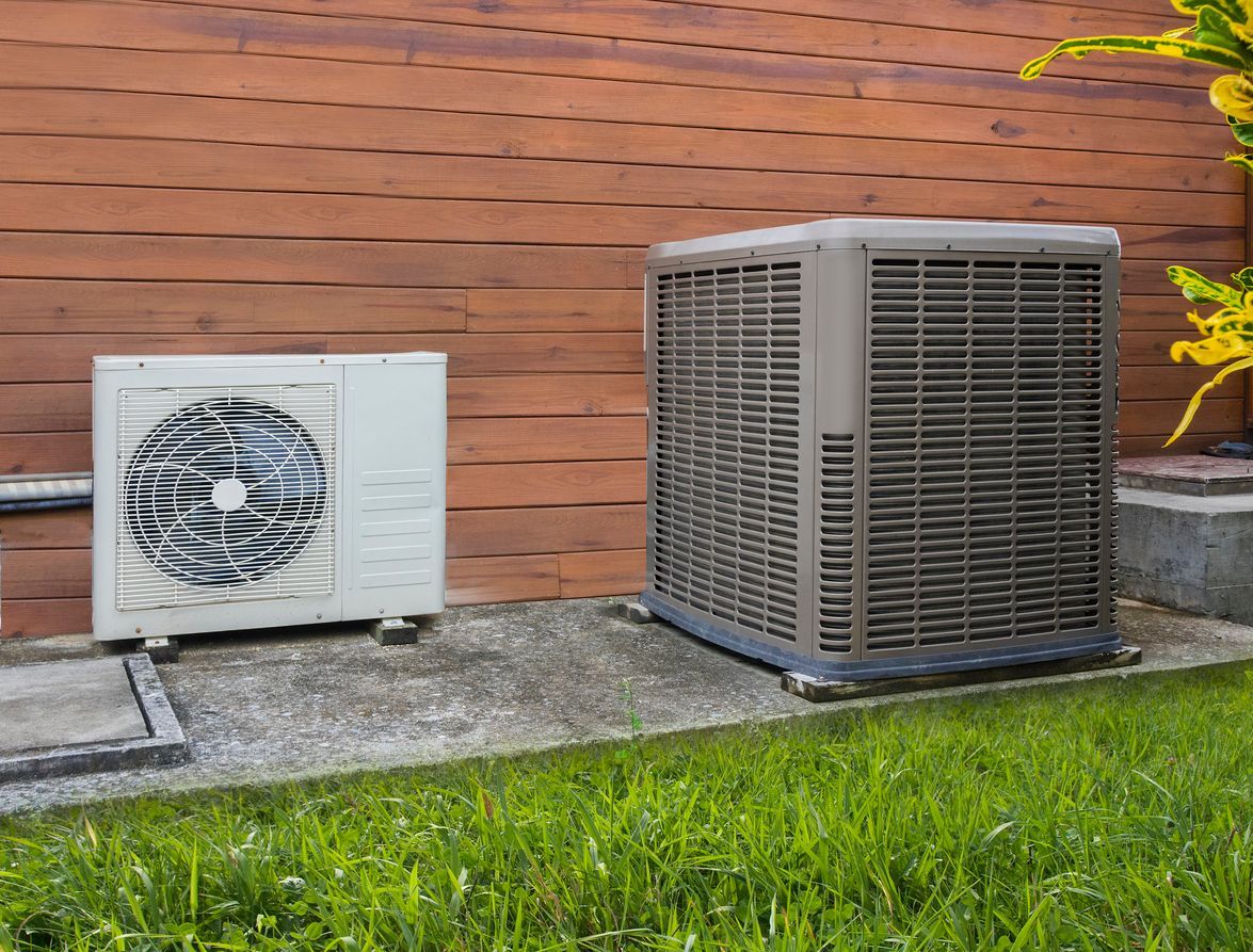 Two HVAC outdoor units, one white and one tan, sit on a concrete slab in front of a horizontal wood slat fence.