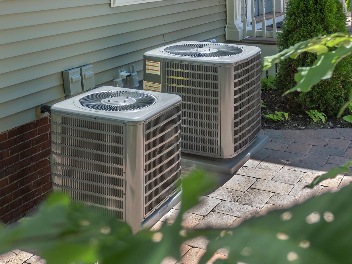 Two metallic residential air conditioning units sit on a paved surface next to a house with beige siding and brick.