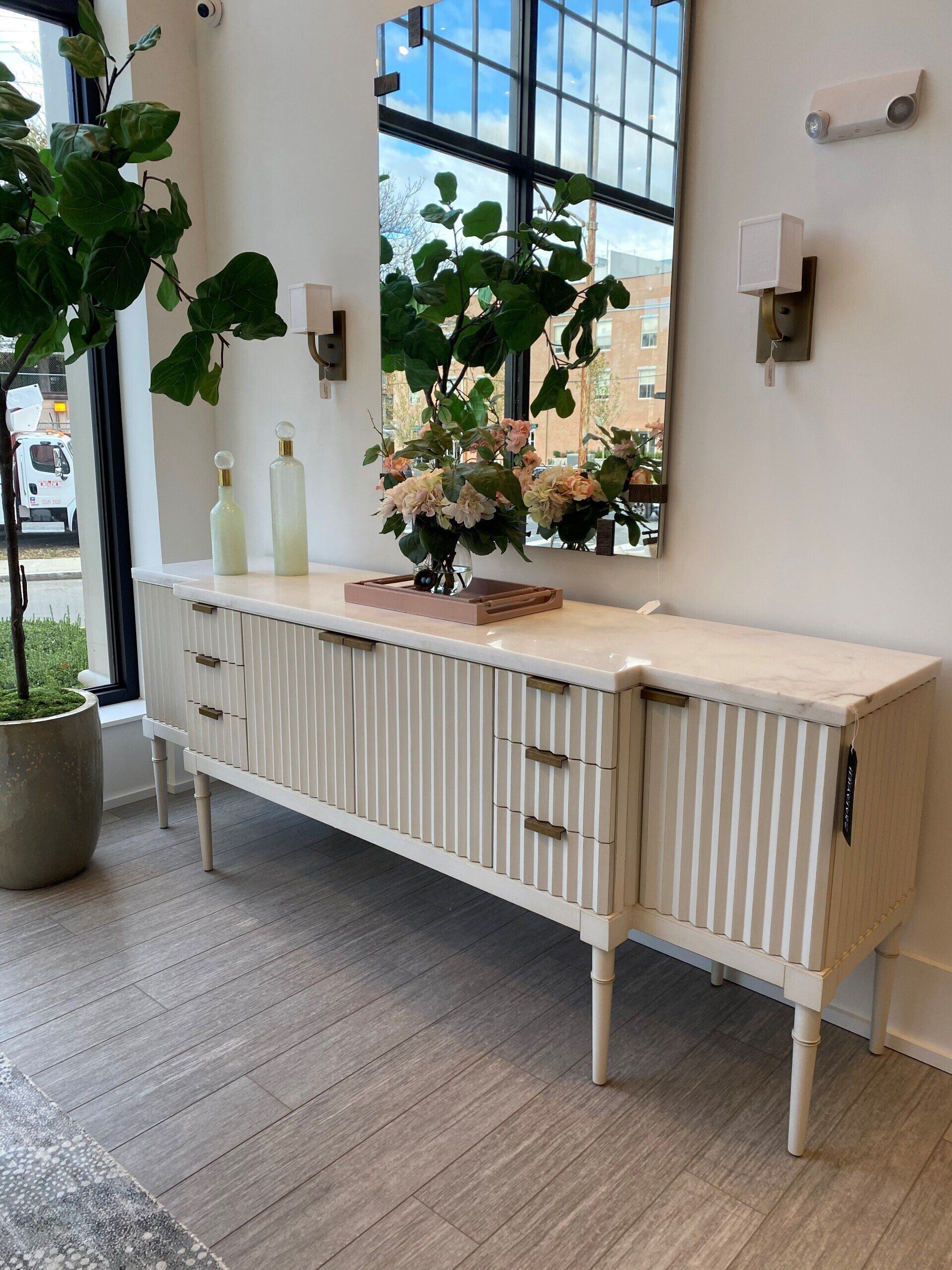 White console table with plants and decor beneath a large window in a bright room White console table with plants and decor beneath a large window in a bright room
