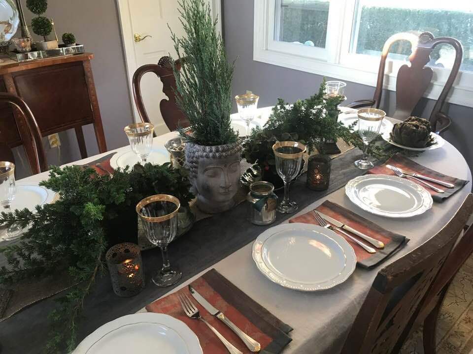 Elegant dining table set with white plates, gold-rimmed glasses, candles, and greenery centerpiece.