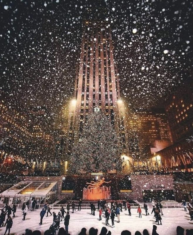 Snowy night at Rockefeller Center with skaters beneath a tall lit skyscraper and falling snow