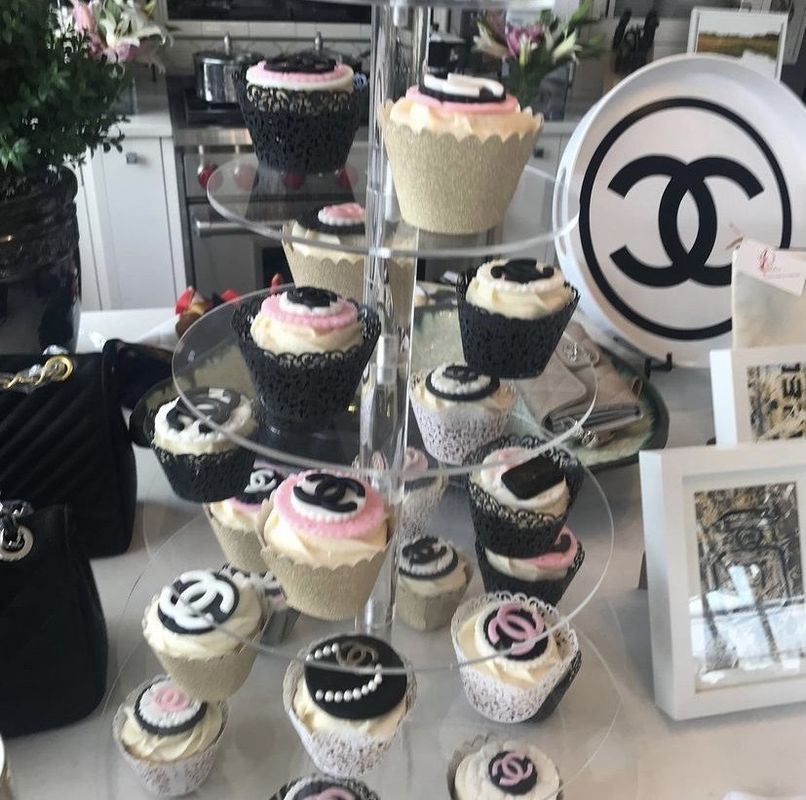 Display of black-and-white cupcakes with pink frosting on tiered trays, beside a large Chanel logo sign