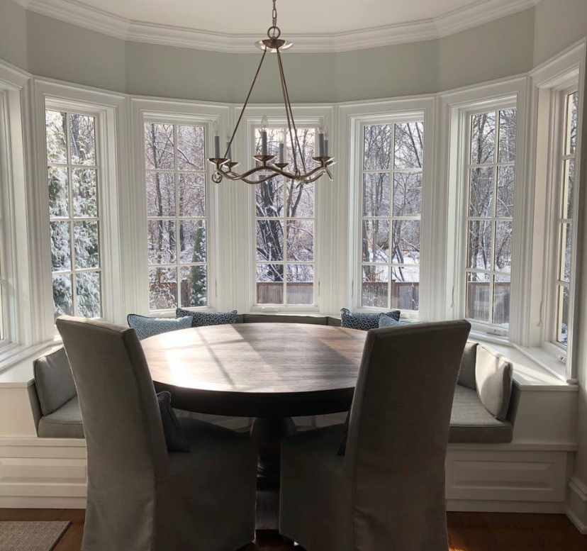 Round dining table and chairs in a bright bay window nook with white trim and hanging chandelier.