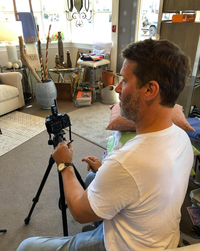 Man holding a camera on a tripod in a cluttered living room, filming toward the window