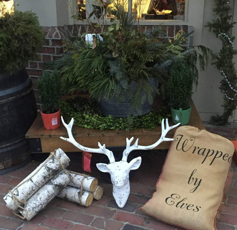 Holiday porch display with white deer head, stacked birch logs, and burlap sack reading “Wrapped by Elves”