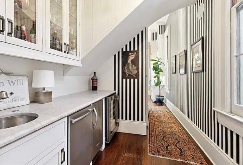 Narrow white hallway with striped walls, wood floor, side counter, mirror, and a runner rug leading to a plant.