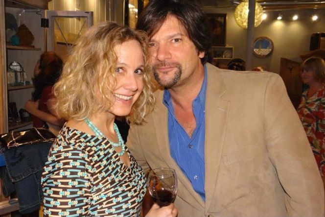 Two smiling people pose together at a warmly lit indoor event, holding drinks.