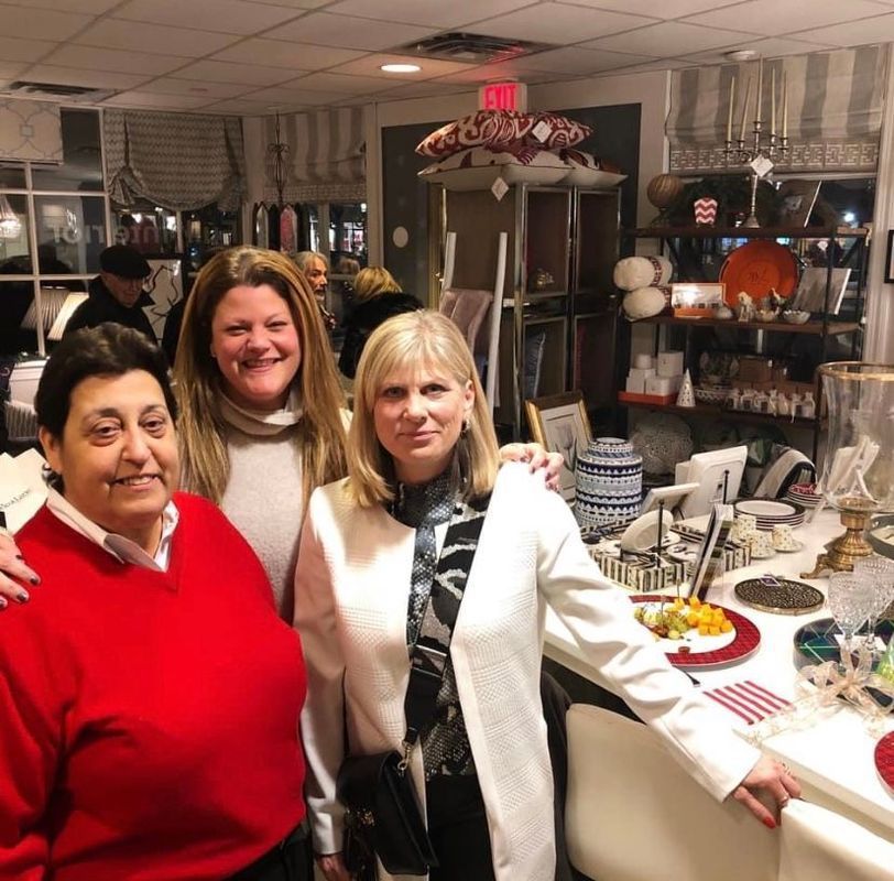 Three women smiling at a home décor shop display table with white, red, and gold items