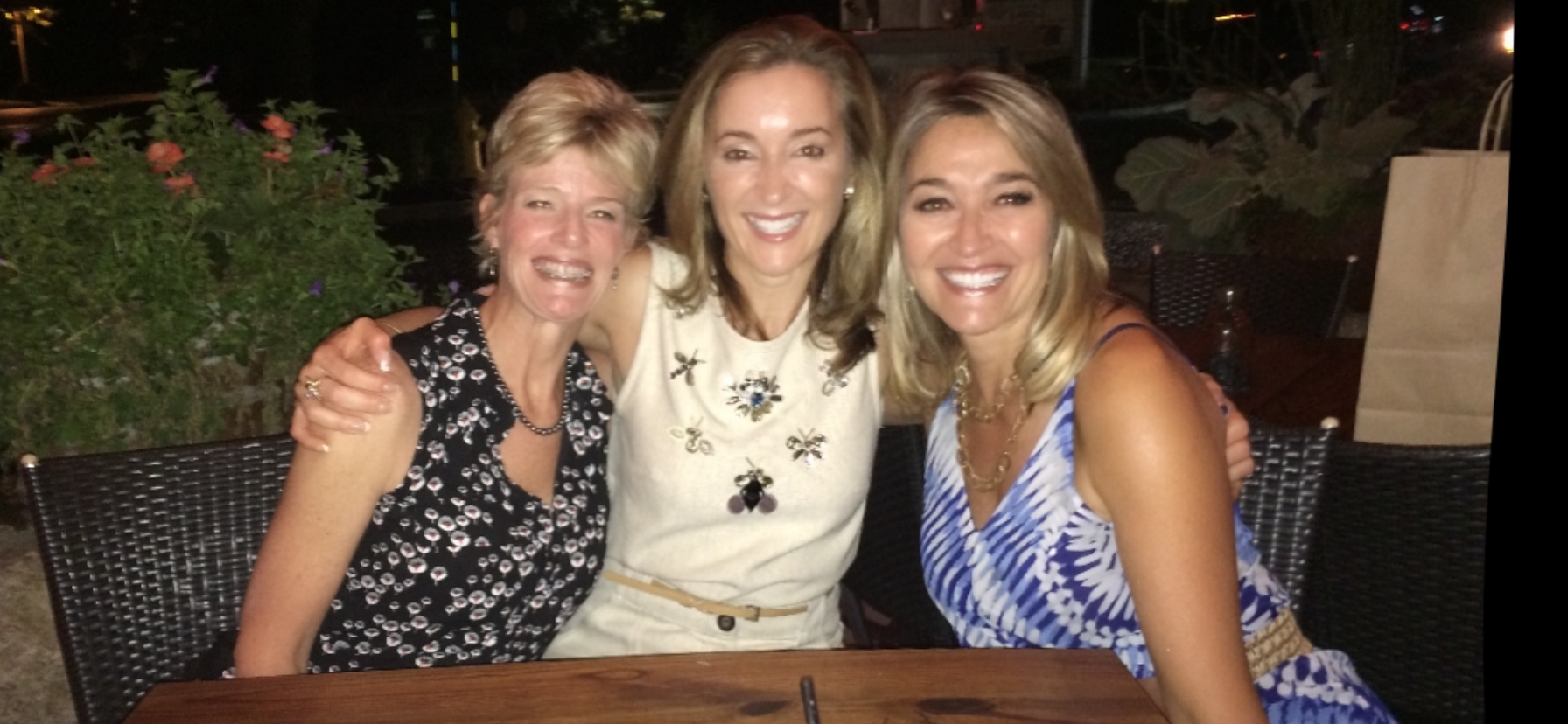 Three women smiling and posing together at an outdoor restaurant table at night.