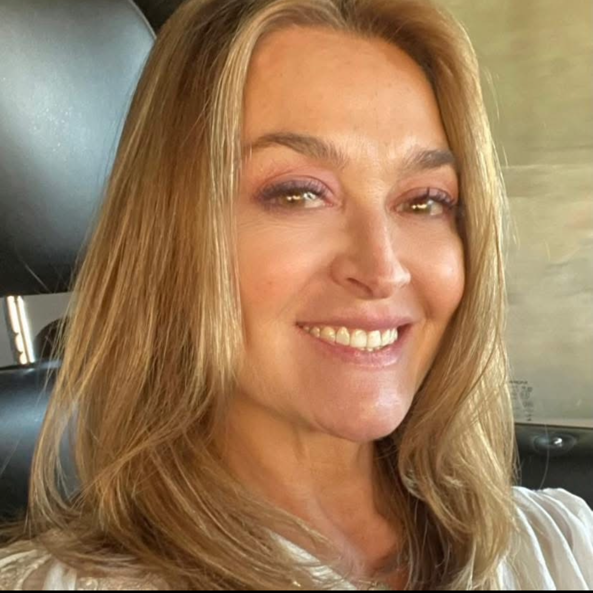 Smiling woman with blonde hair sitting in a car, wearing a white top.