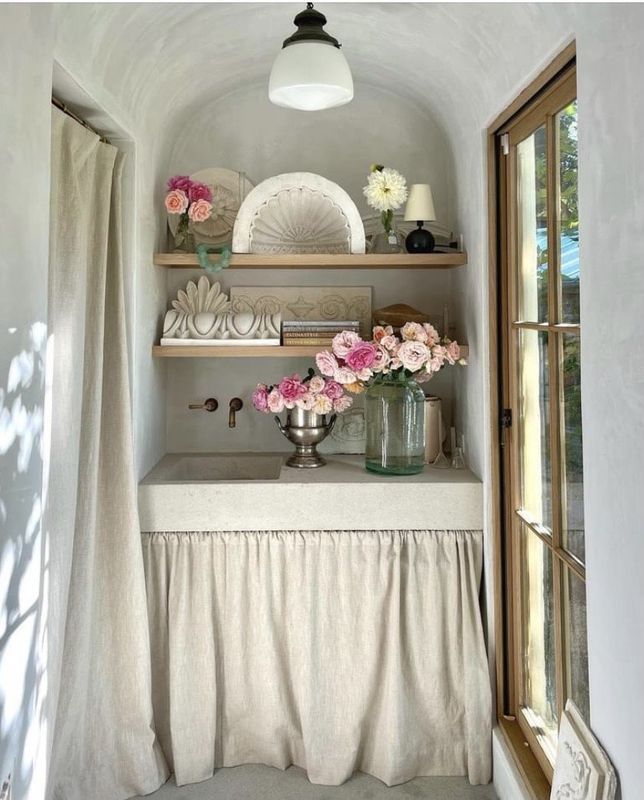 Small alcove with a white curtain, wooden shelves, and pink flowers by a window and glass door.