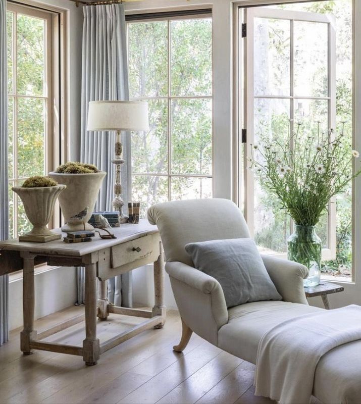 Bright sunlit reading nook with a gray armchair, side table, lamp, and flowers by tall windows
