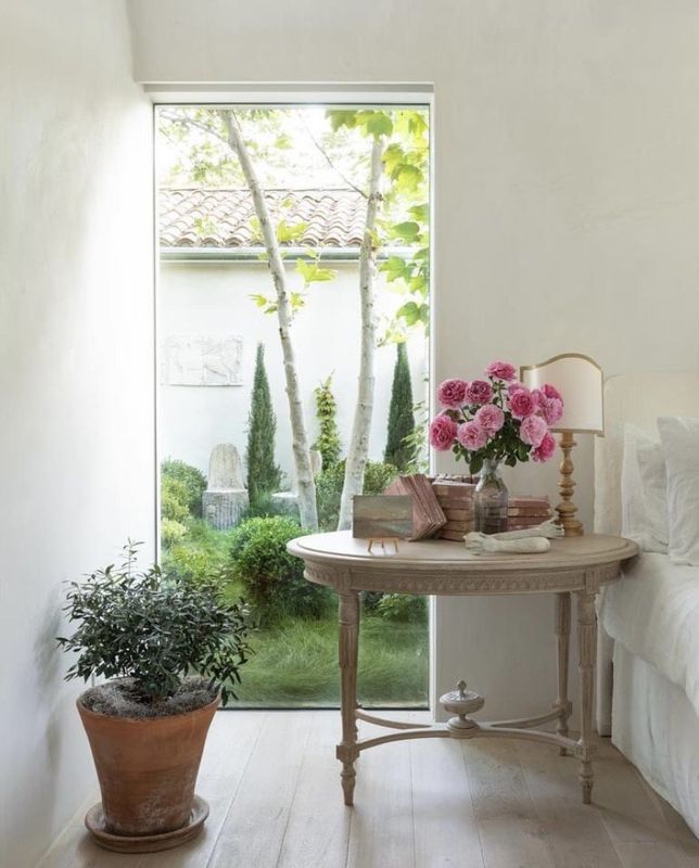 Bright room with a potted plant, round table, pink flowers, and a garden view through a tall window