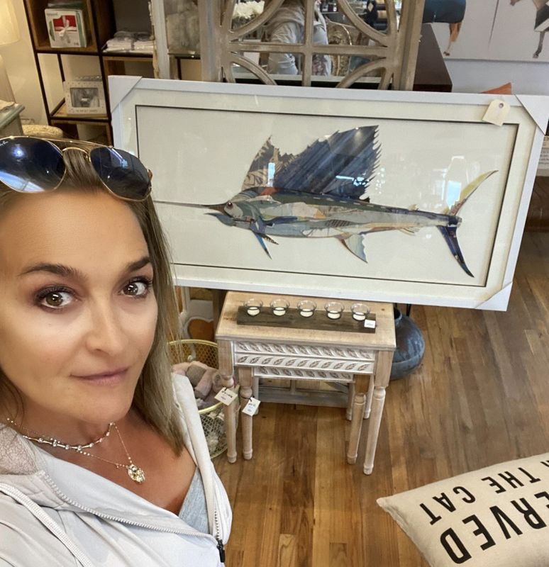 Woman taking a selfie in a shop, with a framed blue fish painting and coastal decor behind her