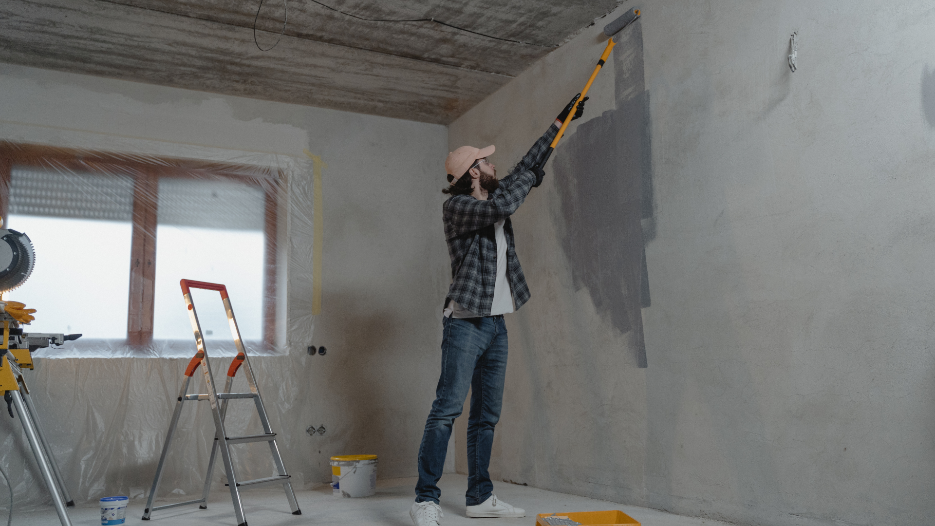 Person painting a gray wall with a roller in a room under renovation, with ladders and tools nearby