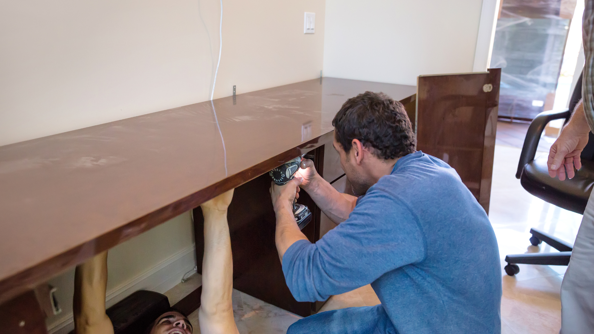 Person kneeling under a desk, using a handheld tool to work on wiring or hardware inside.