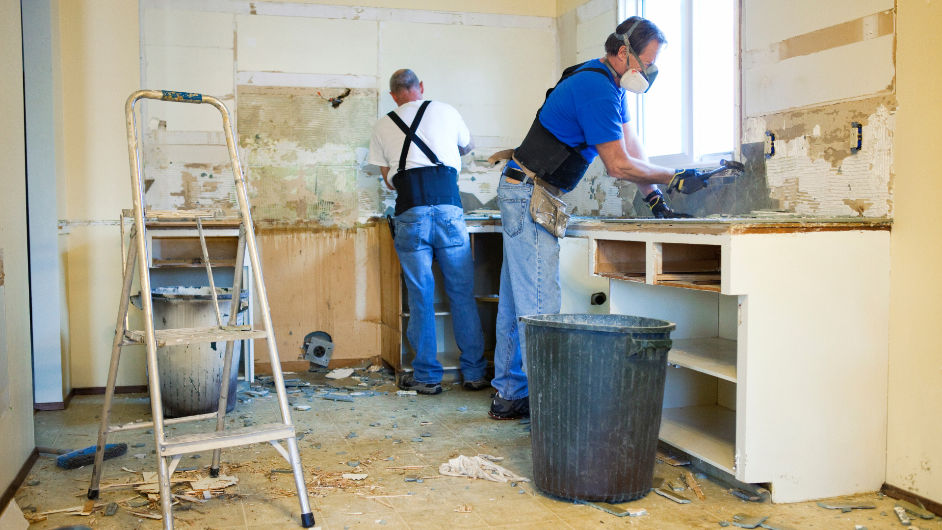 Two workers renovating a damaged kitchen with tools, ladder, and debris on the floor