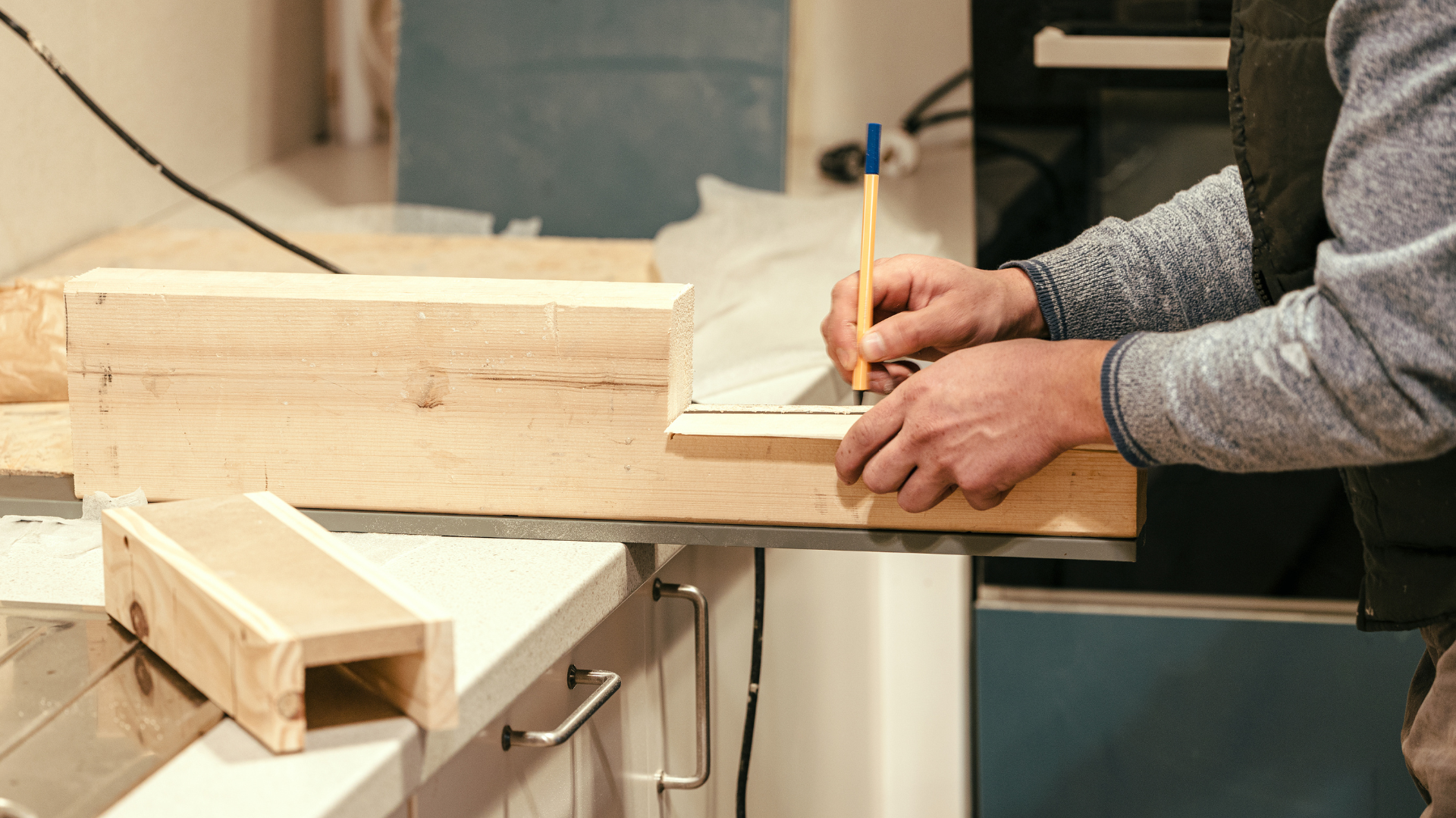 Person measuring wood with a pencil in a workshop beside a miter saw and wooden boards