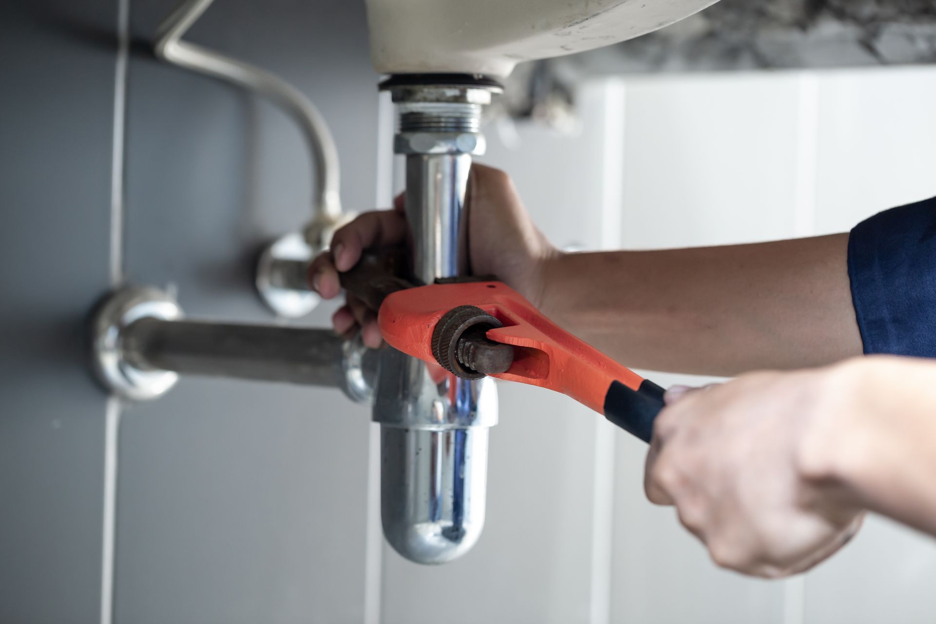 A person uses an orange pipe wrench to tighten a metal p-trap under a sink.