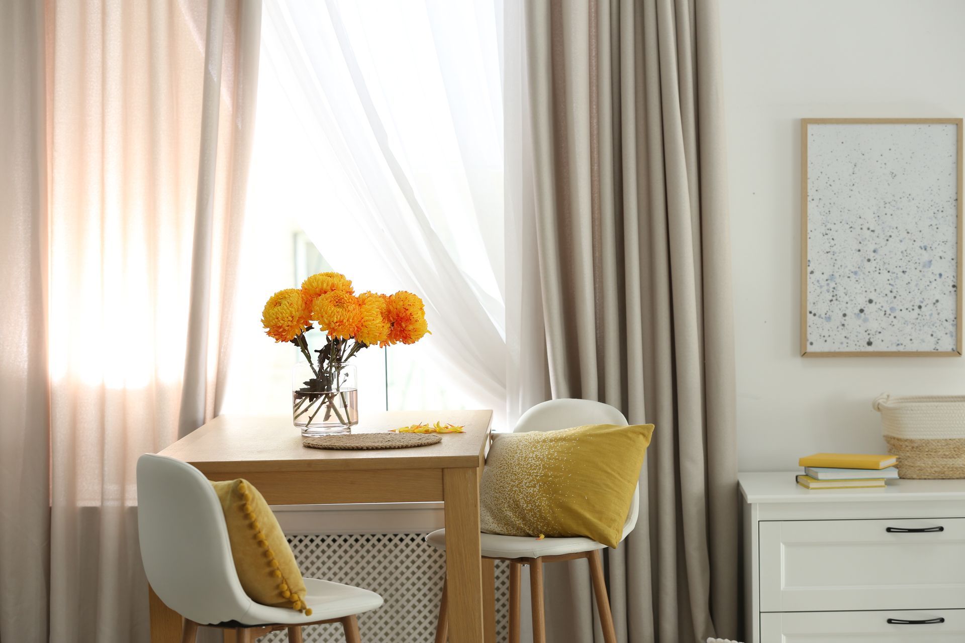 A dining table with yellow flowers, two white chairs with mustard pillows, beige curtains, and a framed art print.