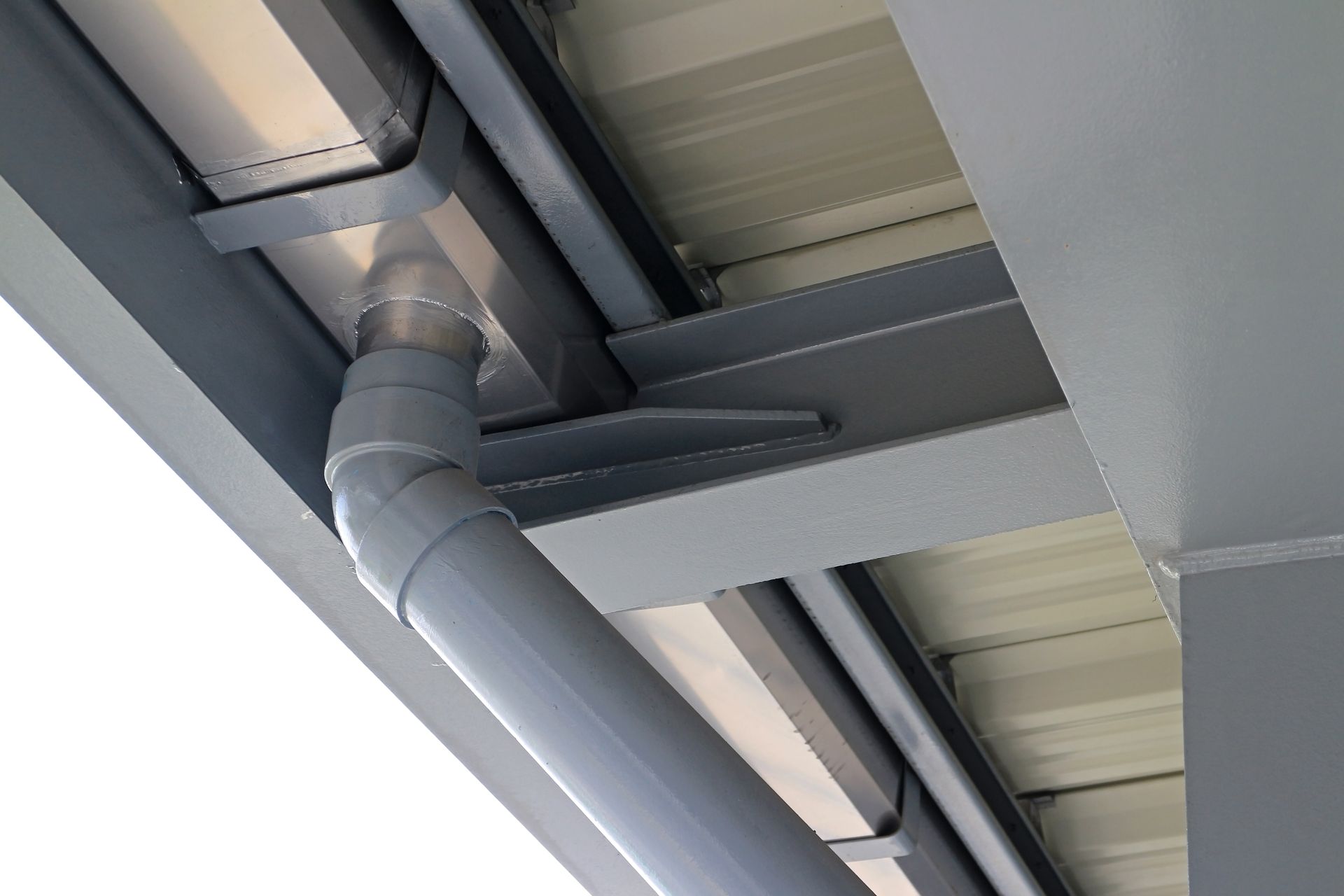 A low-angle view of a gray metal gutter system and downspout attached to the corrugated underside of a building roof.