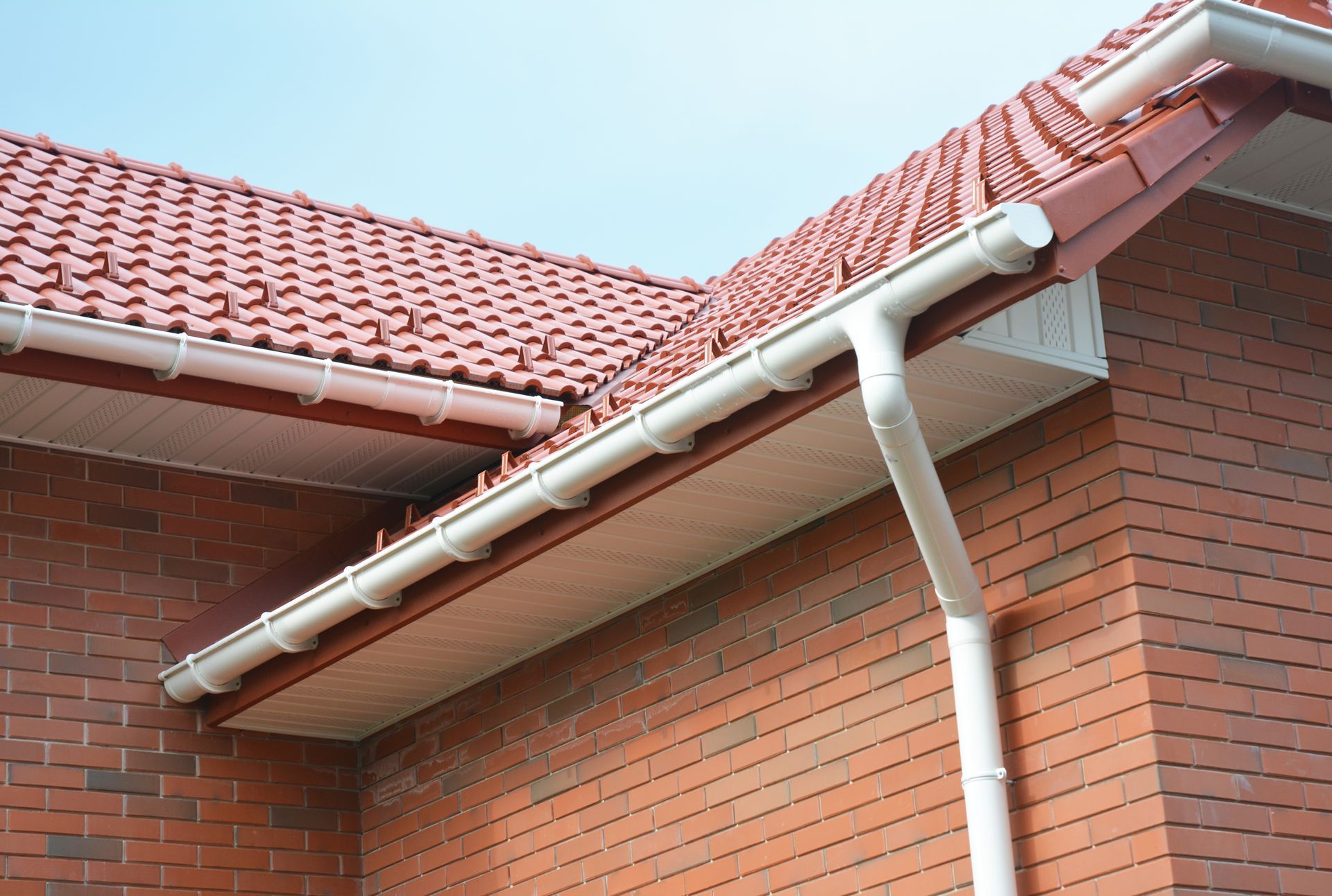 White PVC rain gutters and a downspout attached to the red brick exterior and tiled roofline of a house.